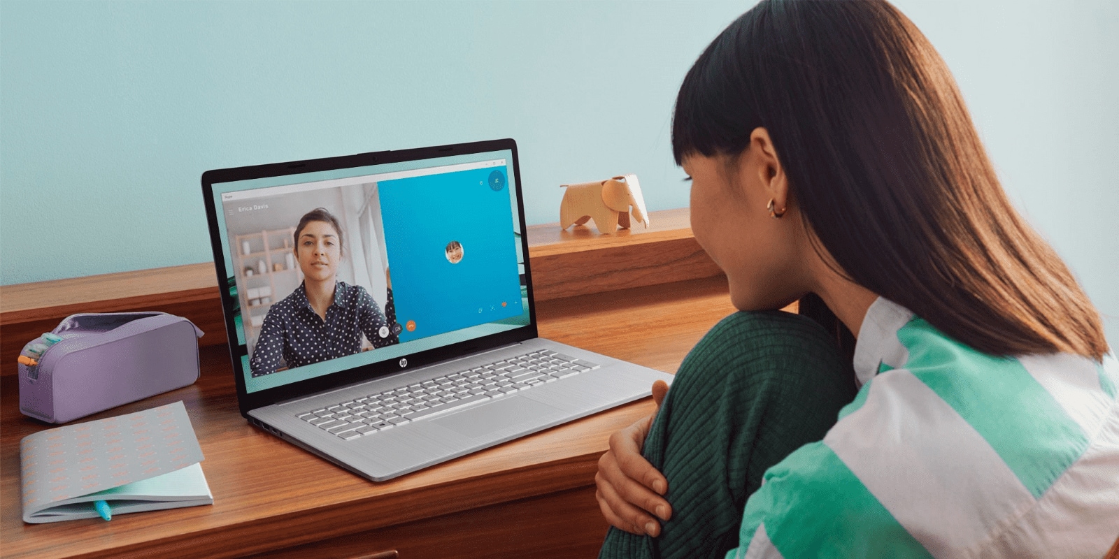 Woman in green sweater on video call with colleague using HP laptop. Wooden desk with notebook and toy elephant. Remote work or online meet.